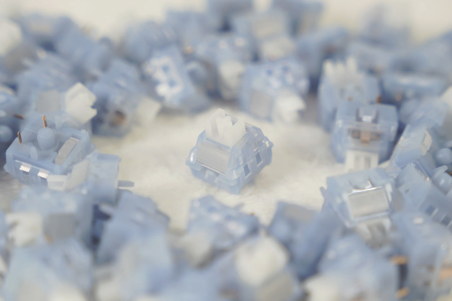 A close-up, shallow depth-of-field shot of multiple light blue and translucent mechanical keyboard switches scattered on a white surface, with one switch in sharp focus in the center.